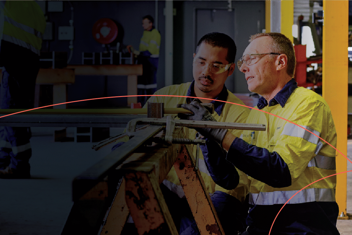 Two workers in yellow safety jackets and goggles focus intently on adjusting machinery in a workshop.
